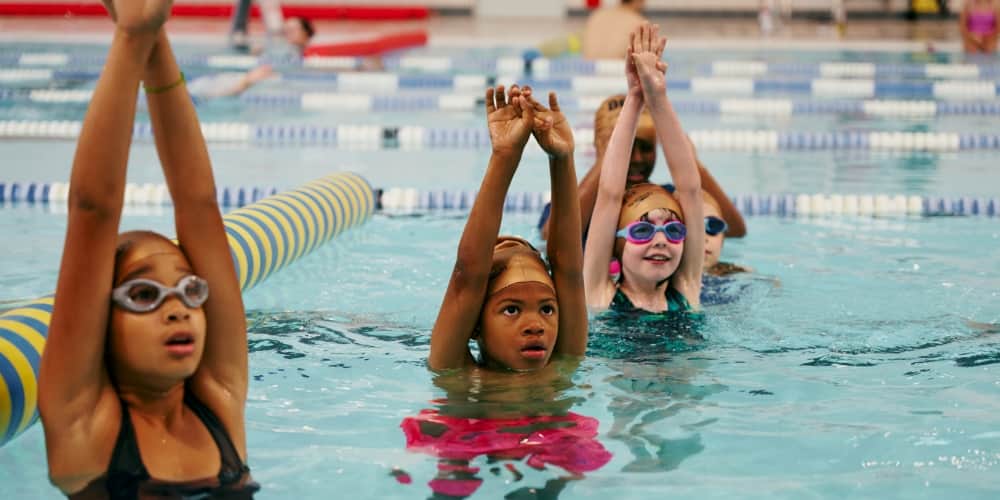 Children raising their arms during a swimming lesson in a pool.